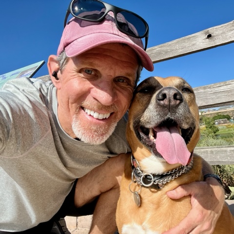 Grey-bearded Steve with a baseball hat and his dog Tesla with her tongue out - both in the sun at a park
