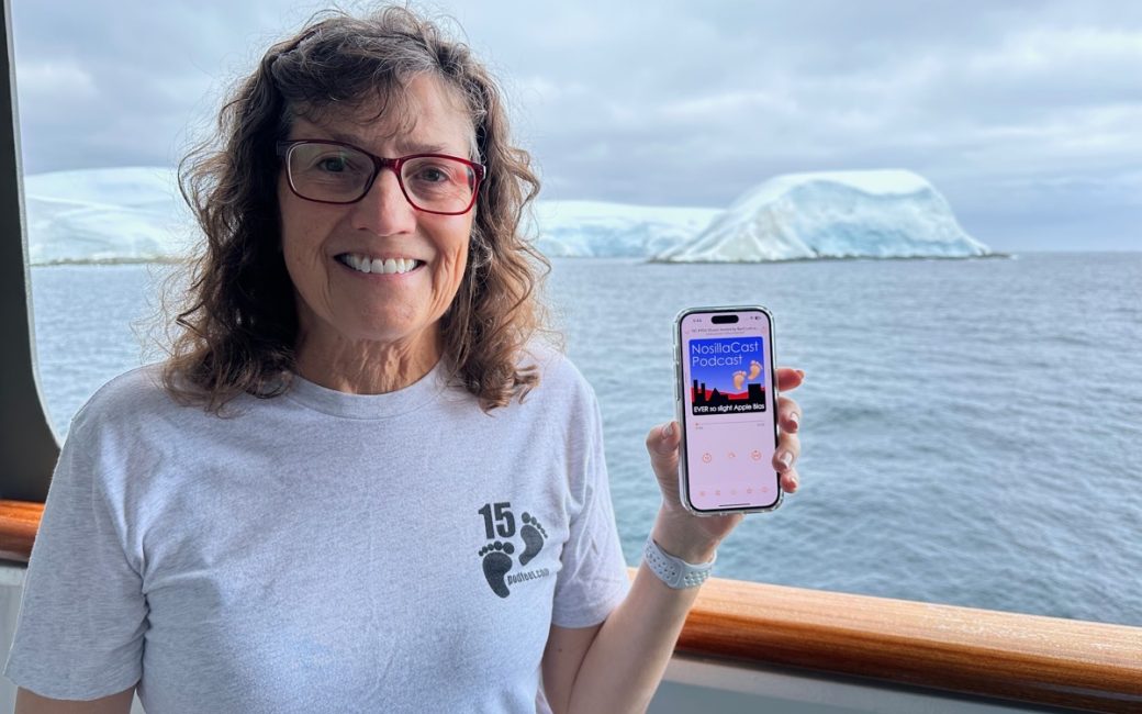 Allison holding an iPhone with the NosillaCast playing. She's wearing a Podfeet t-shirt and Antarctica is in the background from the balcony of the ship