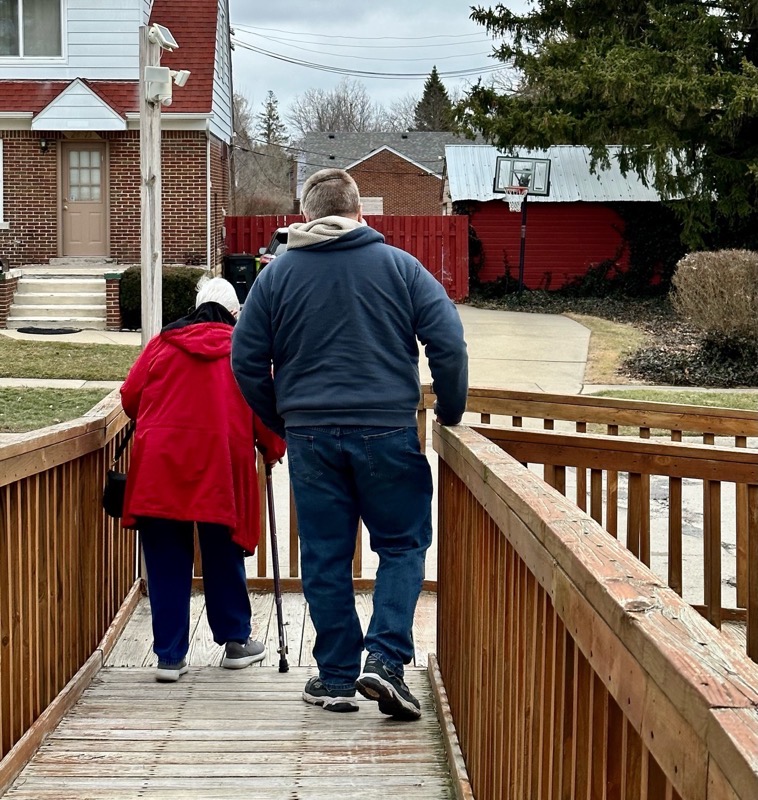 Mom in a red coat and a cane being escorted down a wood ram by a tall man