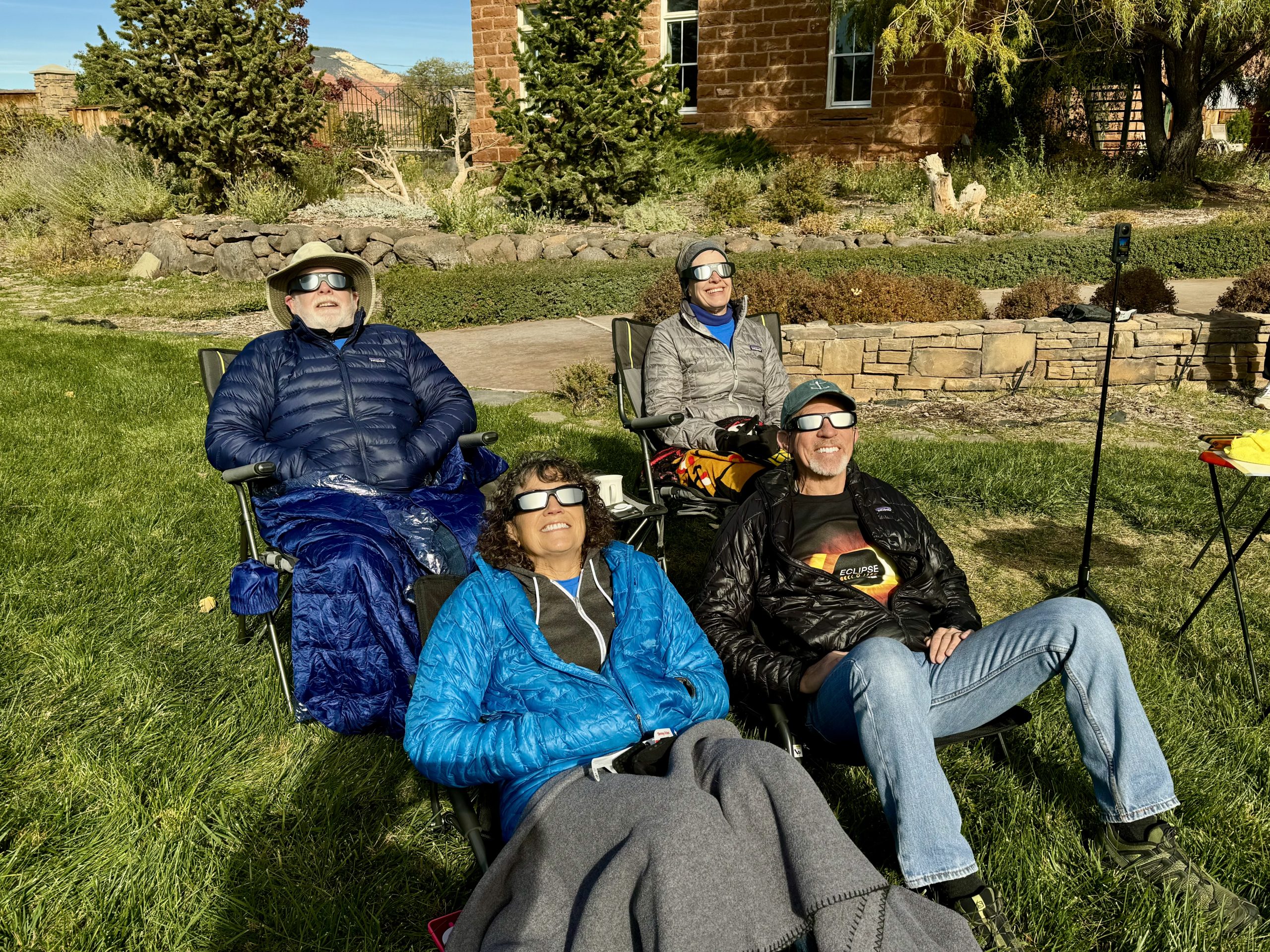 four adults in lawn chairs wearing down jackets with solar glasses on staring at the sun