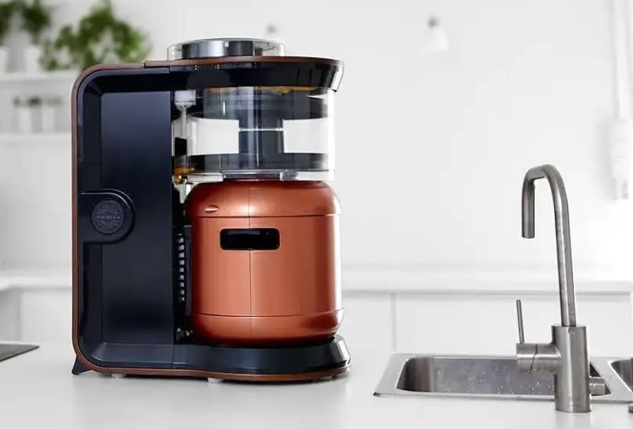 About twice the size of a Keurig coffee maker, the Exobrew device with a bronze keg inside an open black plastic housing sits on a kitchen countertop next to a sink and faucet for scale.