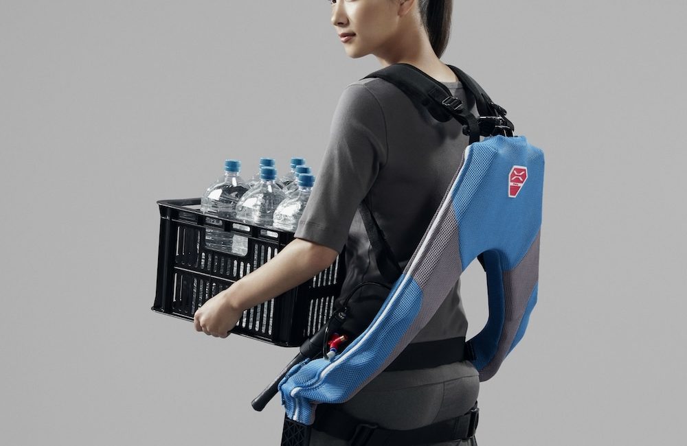 Back quarter-view of a young woman carrying a case of water bottles and wearing the INNOPHYS Muscle Suit. Visible from the rear is the Muscle Suit's exosekleton and its straps to the shoulders, waist, and thighs.