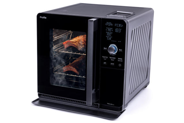 Front view of the GE Smart Home Smoker with a glass door through which smoking meat is visible. The smoker is about the size of a microwave oven and has a display and controls on the front.
