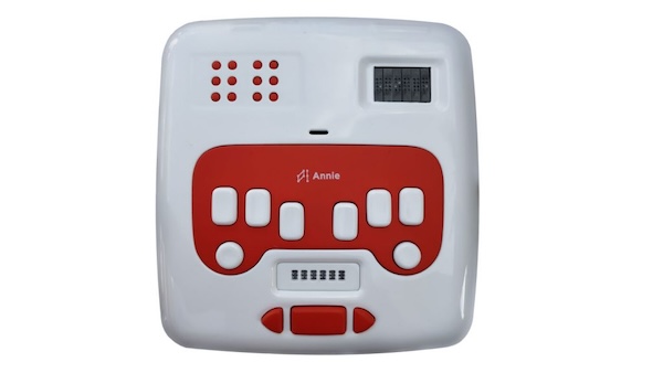 Front view of the handheld Annie device. It's the size of a handheld gaming controller and has a white plastic case with red and white buttons. Visible on the top row of Annie are twin braille cells on the left, speakers in the center, and a standard 6-cell braille display on the right. The middle portion of Annie is composed of a standard braille keyboard with large buttons. Below that is a digital braille slate that lets children input braille characters using a stylus. And finally, at the bottom of Annie are three large buttons for left right/right cursor movement and entry.