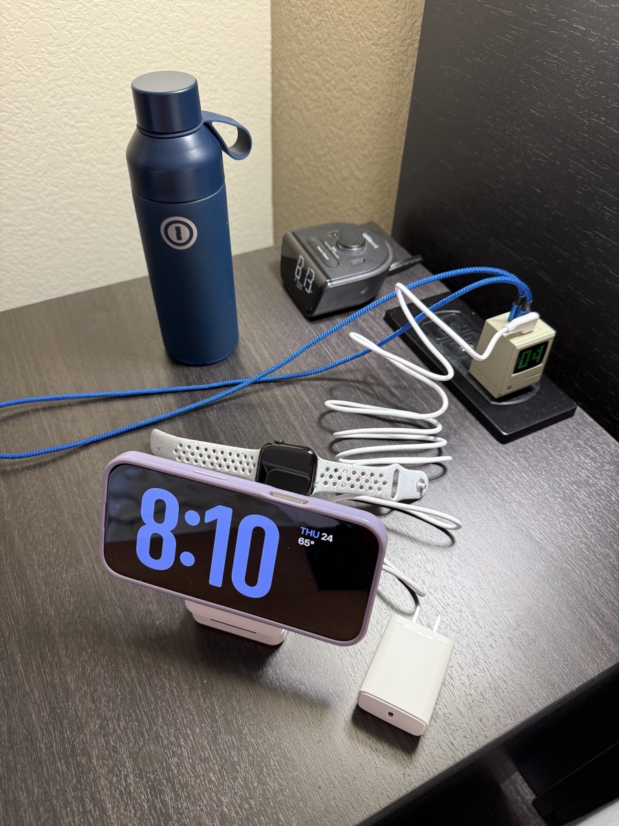 Anker MagGo holding my phone in landscape mode, my watch behind it. it is plugged into the tiny Sharge Retro 67 i am about to explain. you can see the big charger that came with the maggo in the foreground and my 1Password water bottle in the background