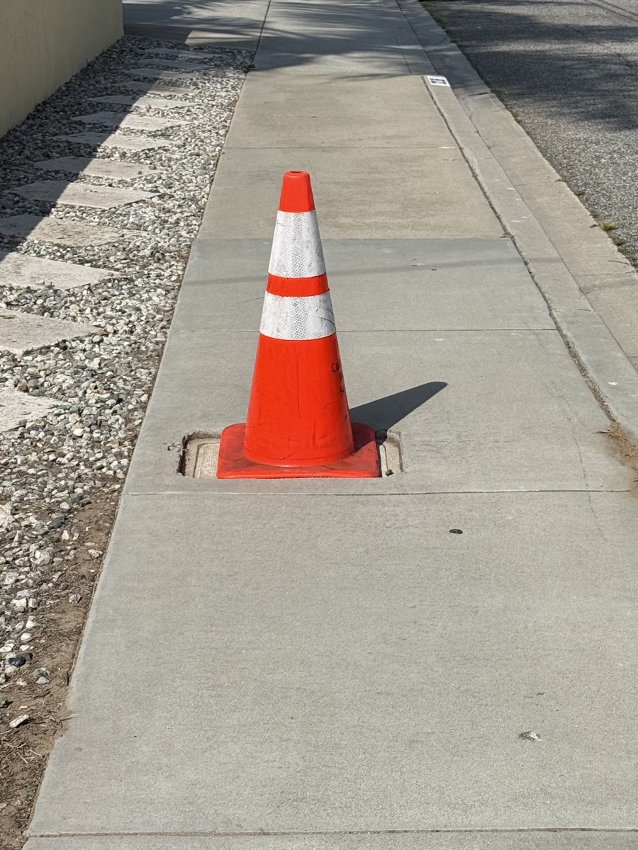 an orange cone sitting in on the water meter cover on a sidewalk