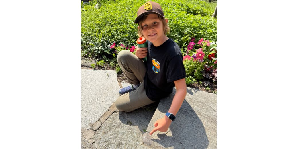 A young boy wearing a baseball cap and a black t-shirt looking up at the camera with a smile. On his left arm is the Apple Watch with a blue band, and he's pointing to his discovery of a Petoskey Stone embedded in the walkway.