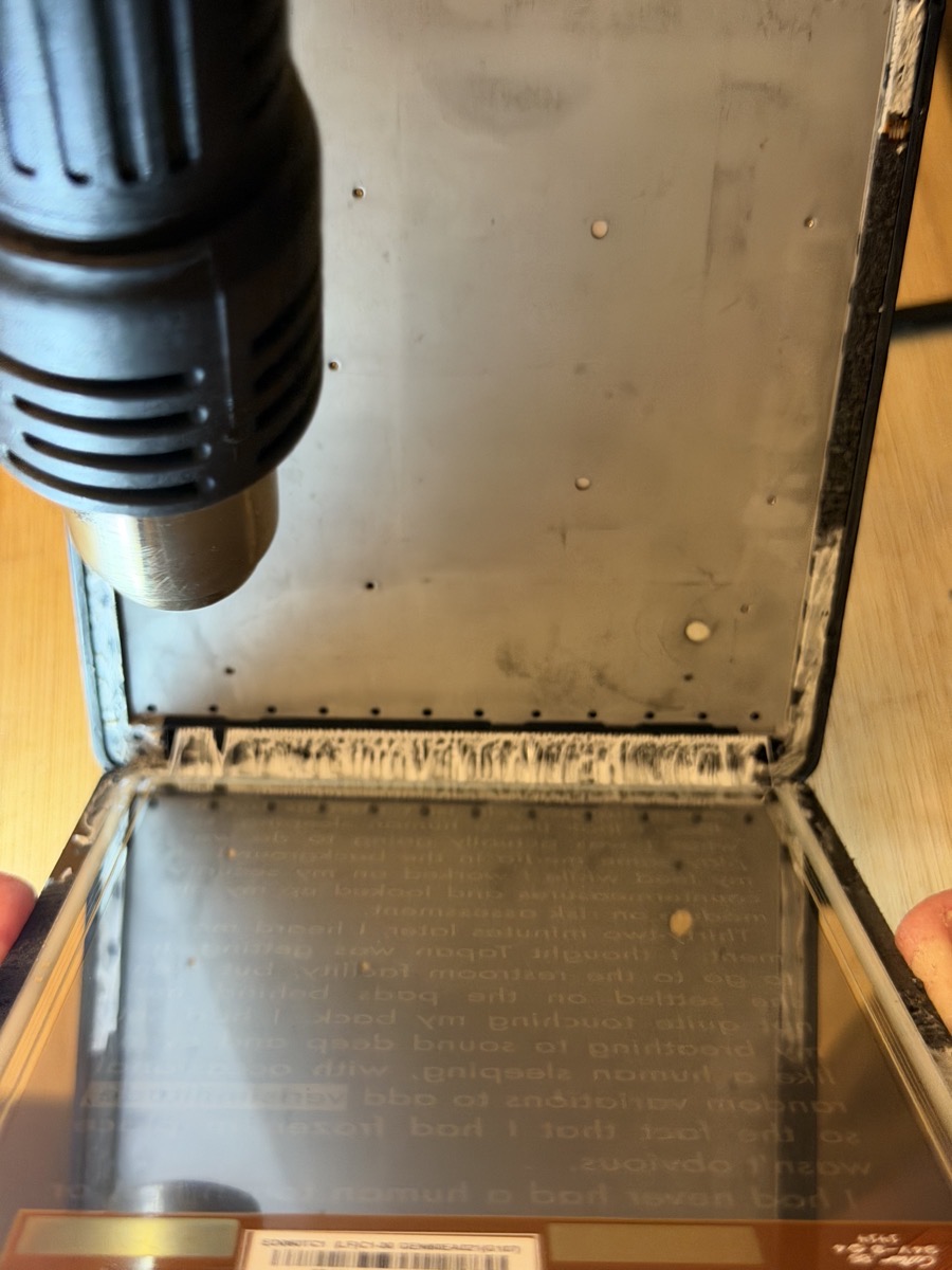 Last edge of adhesive showing between two halves of Kindle and can see text in white on black from the screen including verisimilitude.