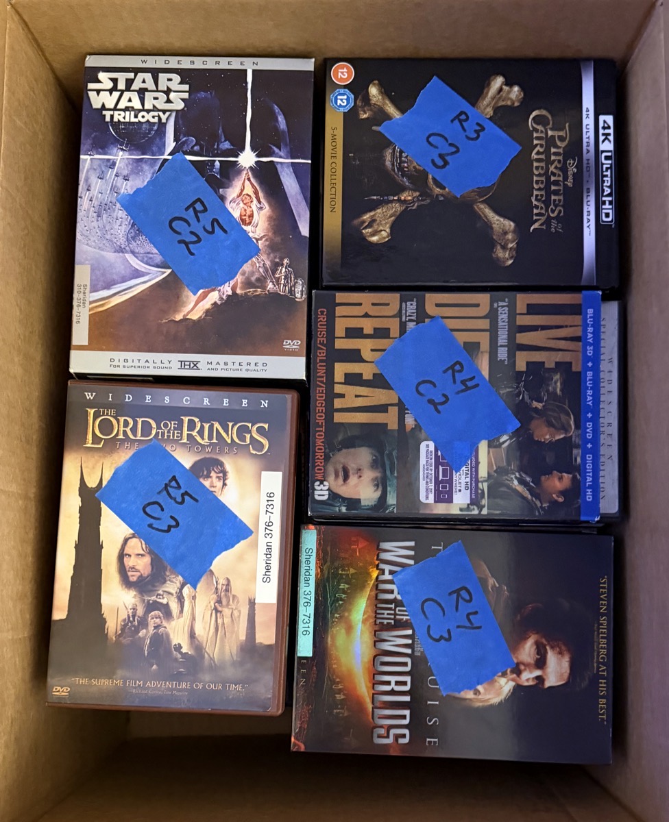 DVDs stacked in a box where each stack is labled with row and column number on blue tape. the movie stacks say star wars, lord of the rings, pirates of the caribbean and two tom cruise stacks