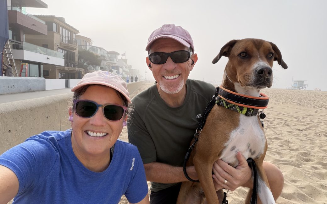 Photo of Allison and Steve kneeling on the beach with Steve holding their dog, Kepler. In the background, the strand can be seen vanishing in the distance on the left and the sandy beach with a foggy sky on the right.