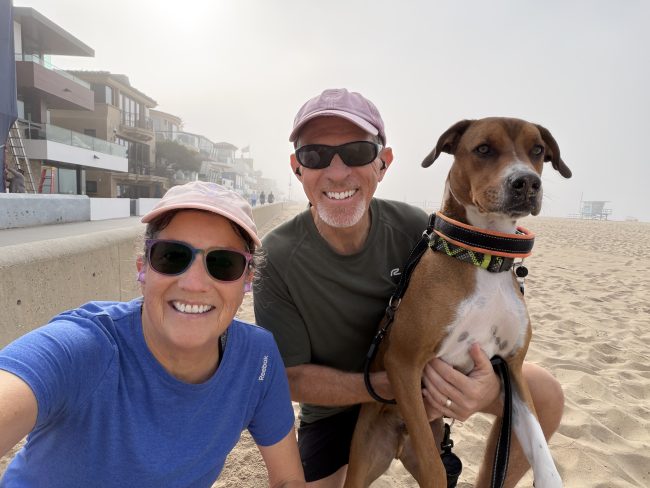 Photo of Allison and Steve kneeling on the beach with Steve holding their dog, Kepler. In the background, the strand can be seen vanishing in the distance on the left and the sandy beach with a foggy sky on the right.