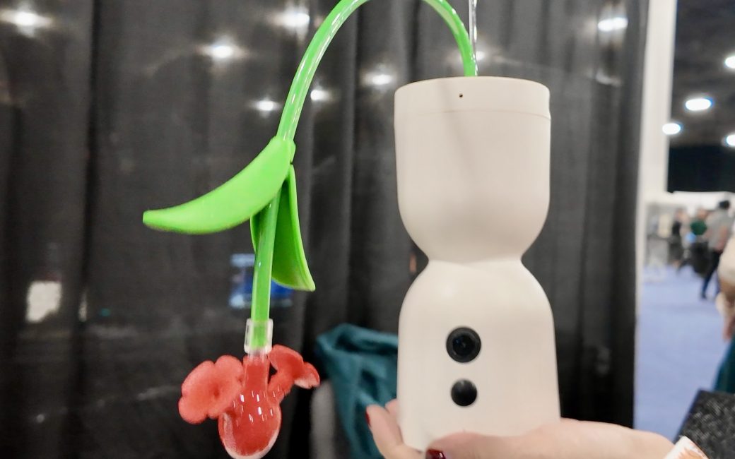 Hand holding a white, cylindrical smart humming bird feeder with two small black camera lenses on the front, a green downward curving tube which models the stem of a flower, and a red flower-shaped dual feeding port. The feeder is displayed indoors at what appears to be a trade show.