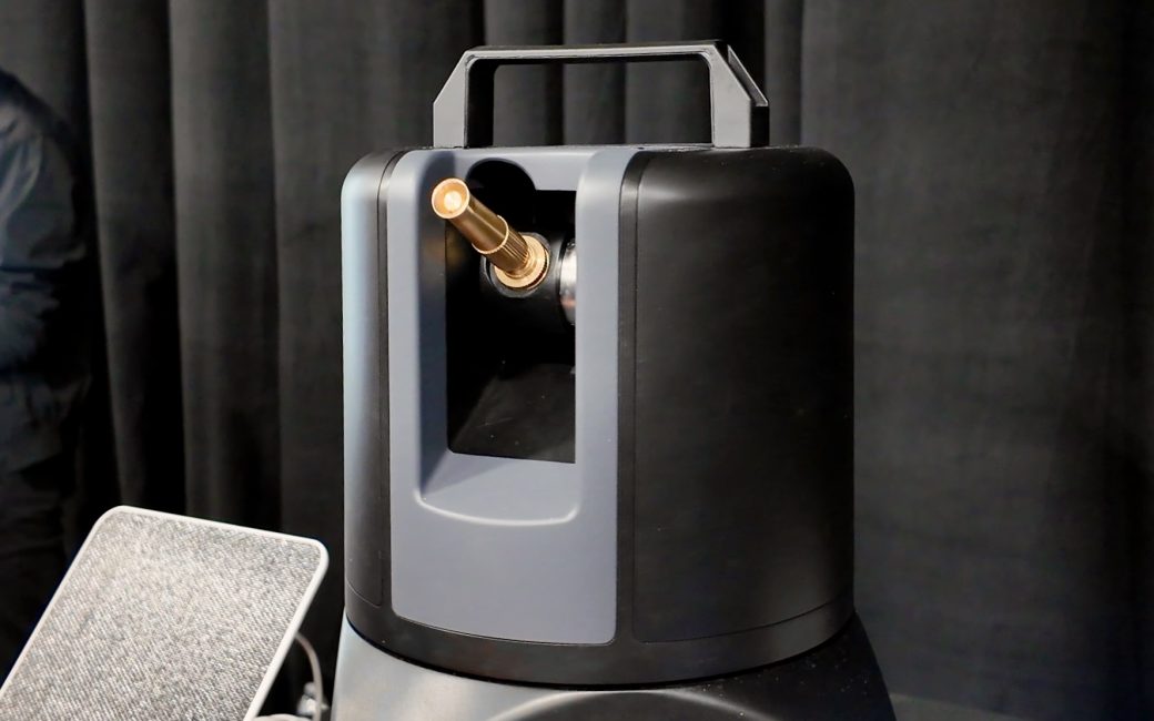 A close-up view of the On Sprinkler, a black and gray cylindrical device with a carrying handle on top. A short brass-colored metal nozzle protrudes from a recessed opening on the front. The device is set against a dark curtain background.