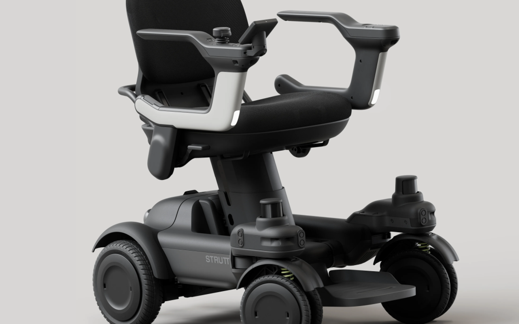 Photo of the Smart EV, a modern four-wheeled mobility scooter with a black padded seat and backrest, elevated armrests with integrated controls, and a compact, sturdy base. The scooter has thick tires, a foot platform, visible suspension components, and a sleek dark gray/black design on a plain light background.