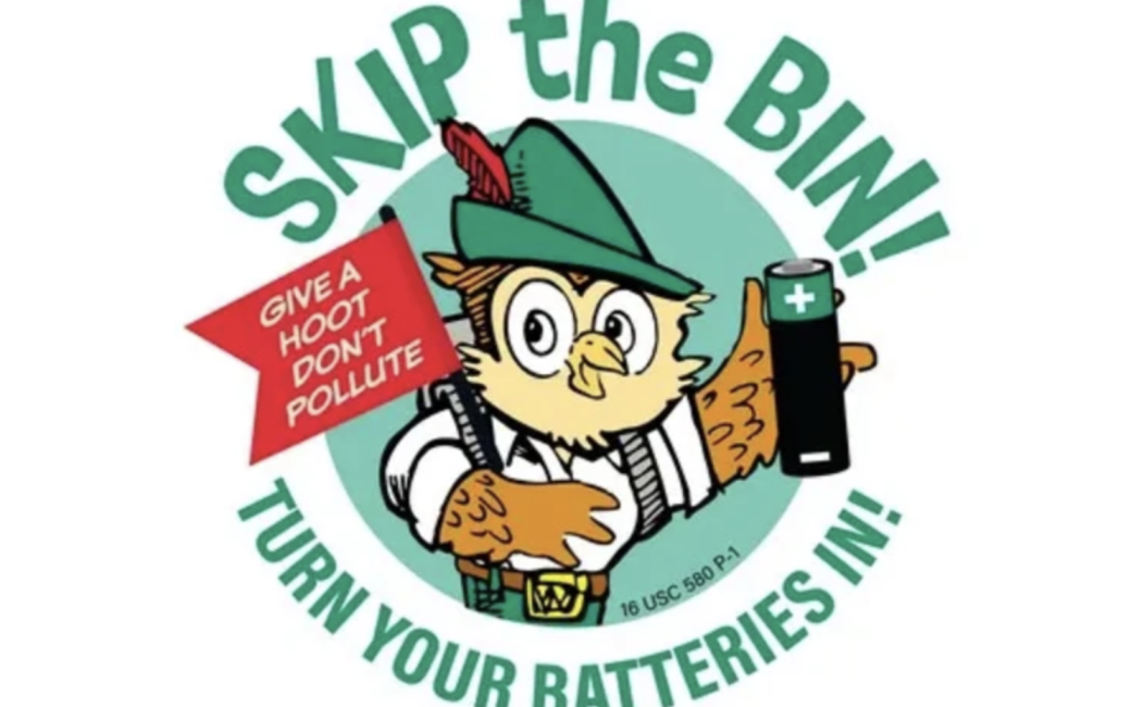 A circular public service graphic featuring Woodsy Owl wearing a green ranger hat with a feather. He is holding a red flag that reads “Give a hoot, don’t pollute” and a battery with a plus sign. Green text around the circle reads “Skip the bin!” at the top and “Turn your batteries in!” at the bottom, on a white background.