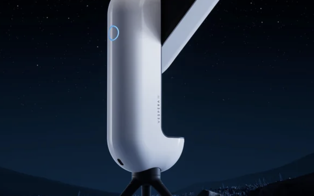 A Vaonis Vespera smart telescope mounted on a three-leg tripod, with a cylindrical white body and an angled telescope section, set outdoors under a star-filled night sky.