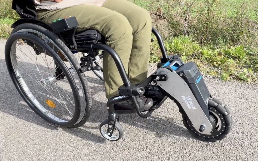 Side view of a manual wheelchair fitted with a WheelMove powered front-wheel attachment, showing the motorized wheel, battery pack, and mounting arms extending from the footrest area as the user sits on a paved path outdoors.