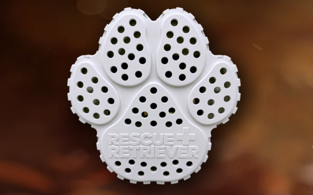 White paw-shaped Rescue Retriever device with perforated holes across each toe and pad, featuring the embossed text “RESCUE RETRIEVER” with a small medical cross symbol. The object has a textured outer edge resembling tire tread and is shown against a blurred warm brown background.