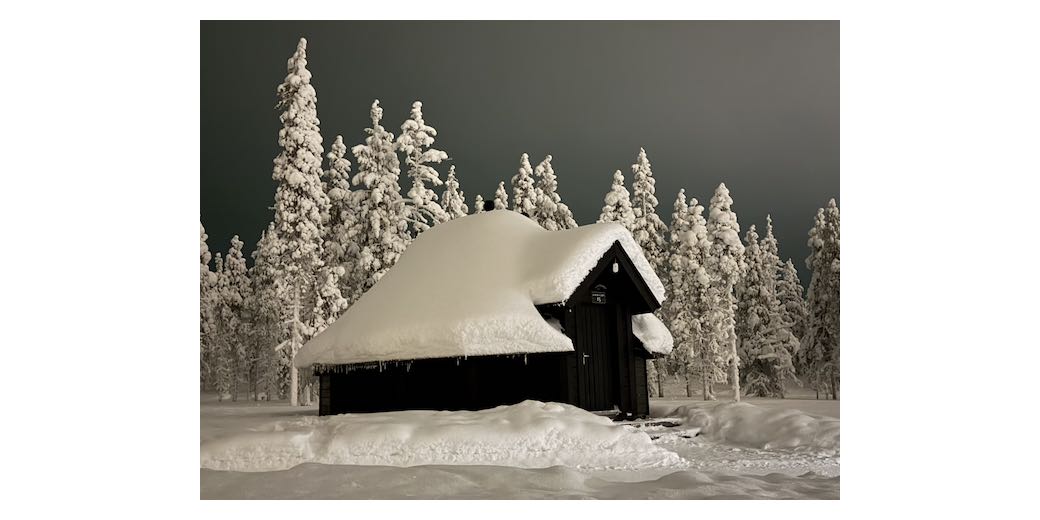 A tiny dark wood hut with 2 feet of snow on top and snow-laden pine trees against a dark grey night sky