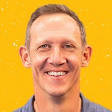 Face shot of Jason Howell with a yellow/orange background. Jason is smiling widely and is wearing a light blue shirt.