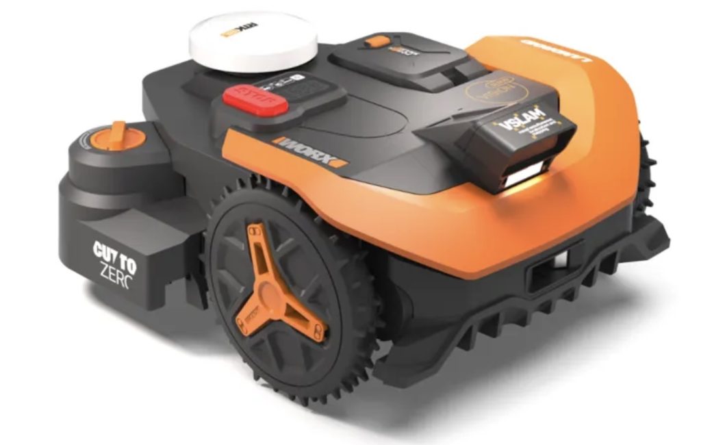 A compact, orange-and-black robotic lawn mower by WORX with large treaded wheels, a top-mounted control panel including a red stop button, and a raised sensor module on top, shown on a plain light background.