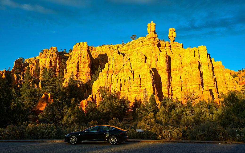 Ron Model S in Red Canyon Southern Utah. the car is super shiny and beautiful and the backdrop is dramatic cliff rocks bathed in an orange glow from the sunset with a rich blue sky behind.
