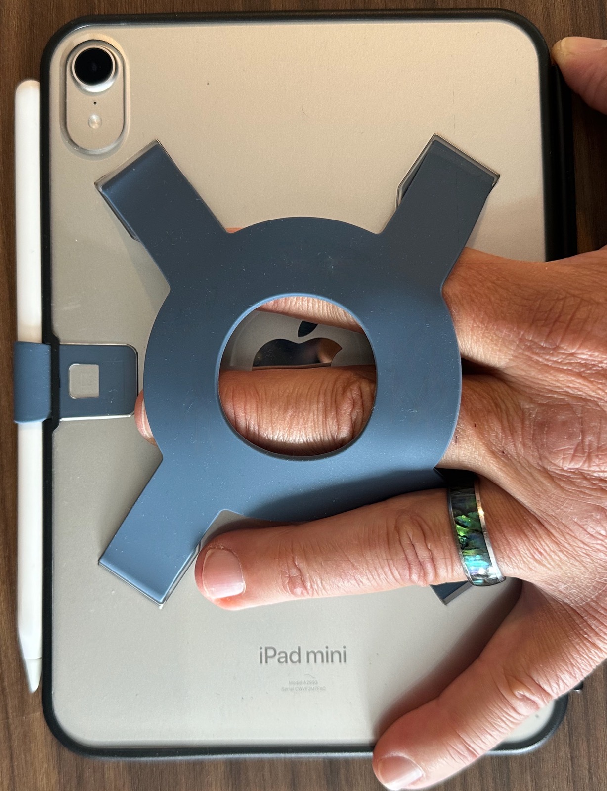 Holding-from-the-side-full.jpeg Back of the iPad mini in the Strapsicle case. A blue-gray silicone harness in an X shape, with a central ring, is bonded to the back of the case. A hand is coming in from the right side of the image, with the first two fingers through the harness.