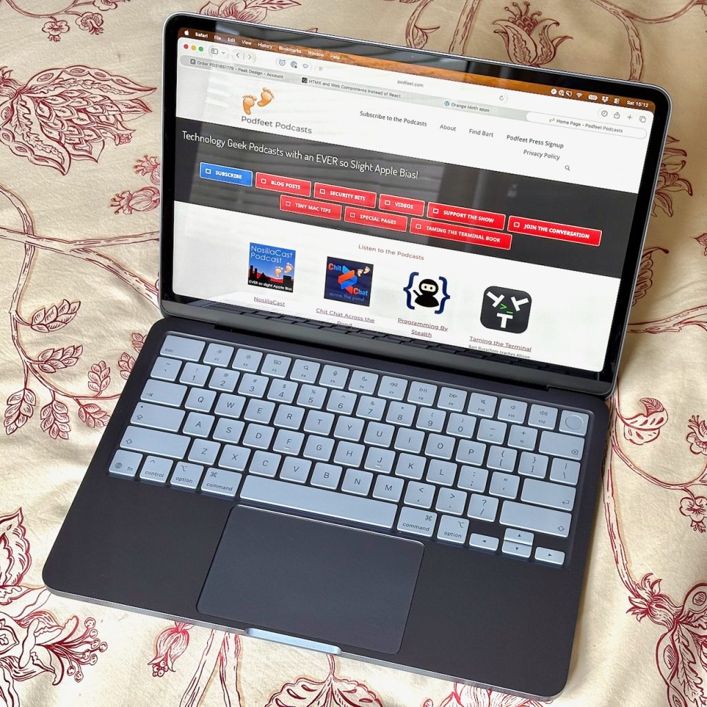 A photo looking down on an Indigo coloured MacBook Neo. It is open with the screen a little past the vertical and it is sitting bed linen which is beige with maroon line drawings of flowers. On the screen of the laptop is the podfeet.com web site. Of course.