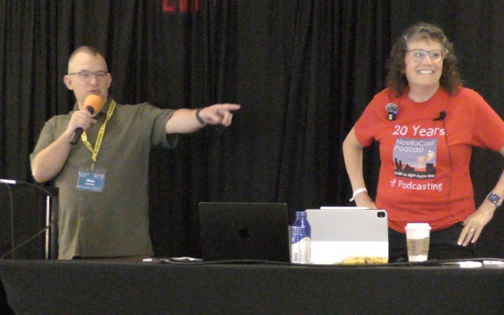 Brian Henson introduces Allison Sheridan at the Macstock conference. Brian speaks into a microphone while pointing toward the audience, while Allison smiles beside a table with laptops, an iPad, a water bottle, and a coffee cup. She is wearing a red T-shirt that reads, "NosillaCast Podcast / 20 Years of Podcasting."