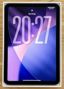A photo of my iPad mini sitting in its box, ready for sale, but the screen is on showing the lock screen with default wallpaper of purples and blues and a large pale, glassy time, which reads 20:27. The box is sitting on a wooden desk.