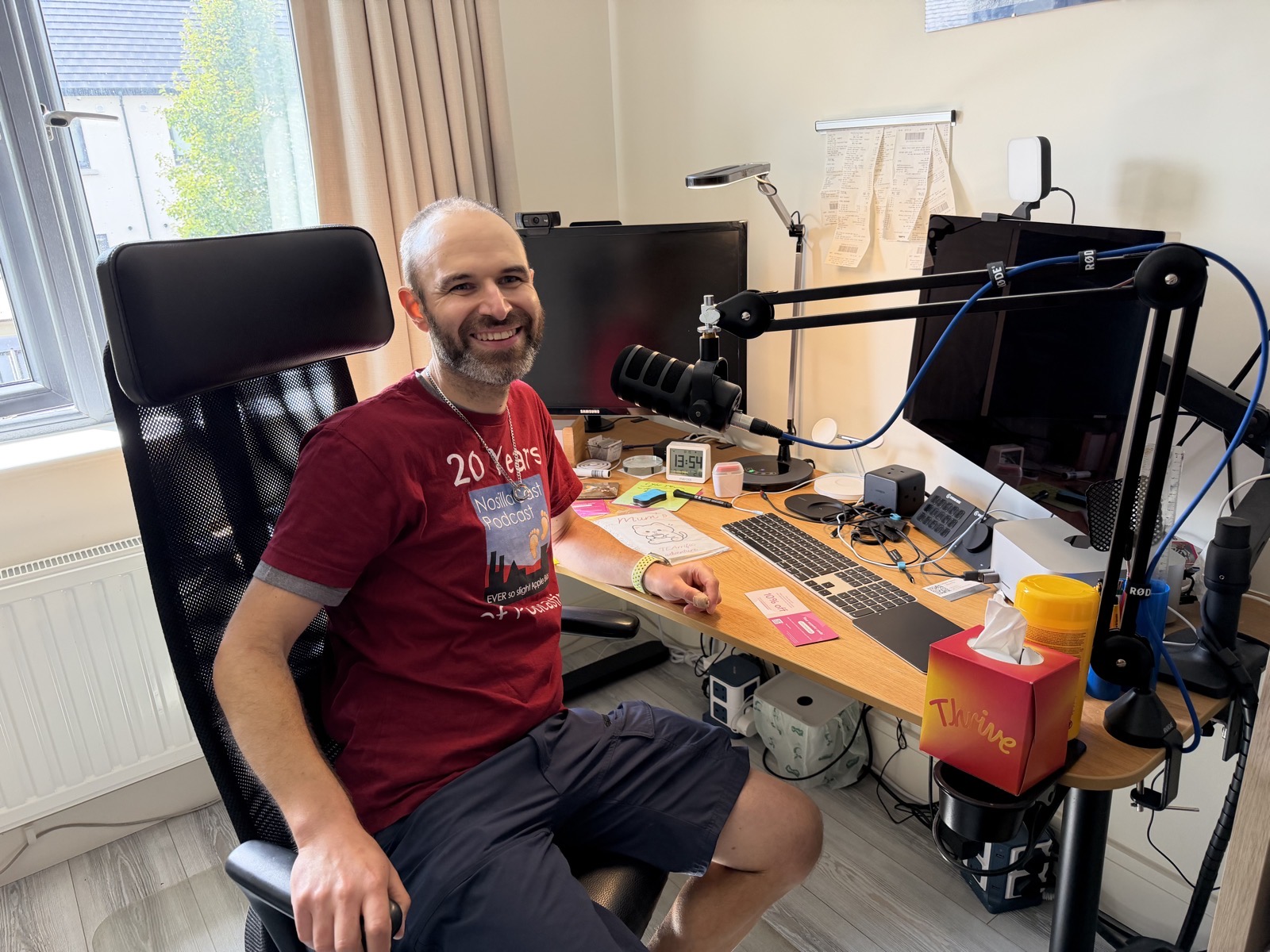 #title# Bart sitting in his chair in his studio wearing a 20 year NosillaCast shirt, with a mic in front of him and his Mac Studio with Studio display