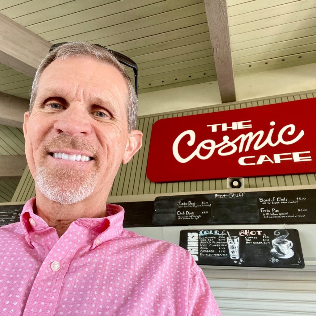 Allison, Steve & Kepler at the Beach Smiling man in a pink patterned shirt posing for a selfie in front of a “The Cosmic Cafe” sign and menu boards.