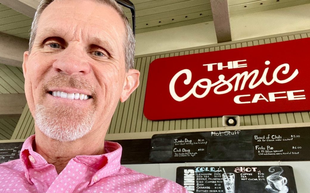 Smiling man in a pink patterned shirt posing for a selfie in front of a “The Cosmic Cafe” sign and menu boards.